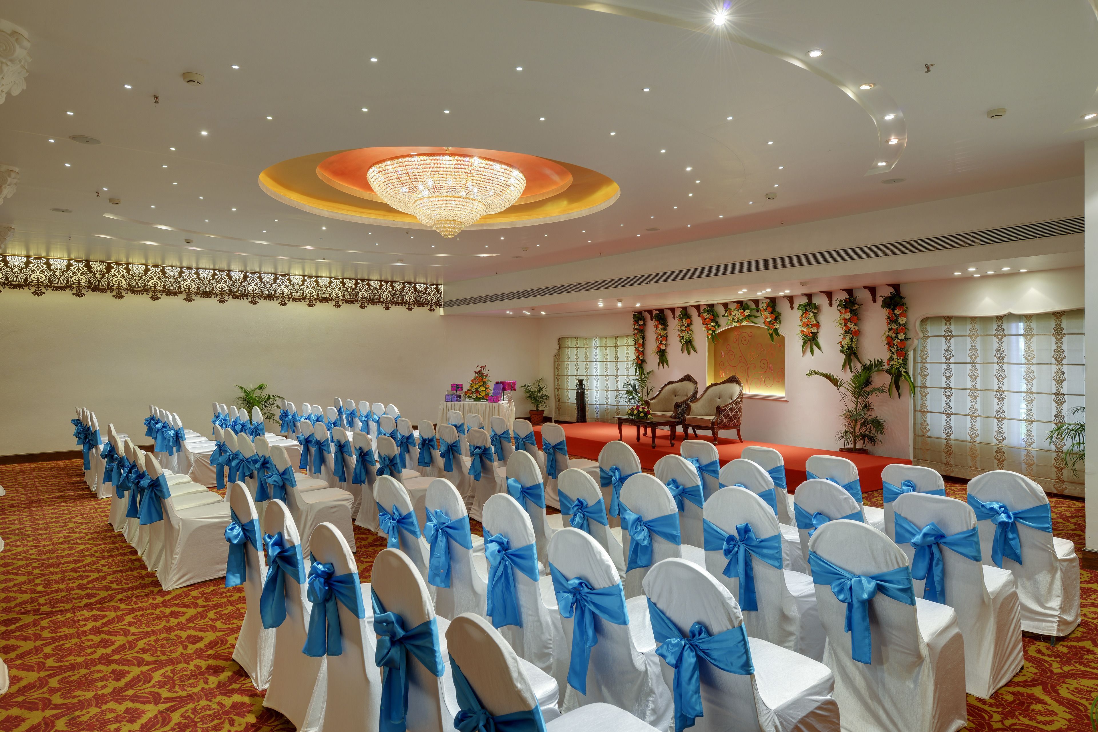 A banquet hall with rows of white chairs with blue sashes, a stage with floral decorations, and a chandelier.