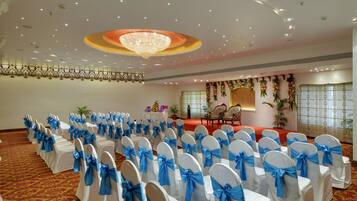 A banquet hall with rows of white chairs with blue sashes, a stage with floral decorations, and a chandelier.