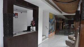 A reception area with a dark reception desk, a vase of pink flowers, and a large colorful mural on the wall. - Hotel Palace Heights (New Delhi)