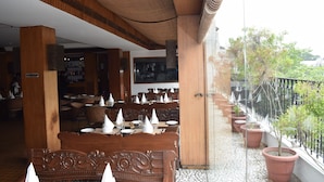 An indoor dining area with wooden tables and chairs, a no smoking sign, and potted plants along a balcony railing. - Hotel Palace Heights (New Delhi)