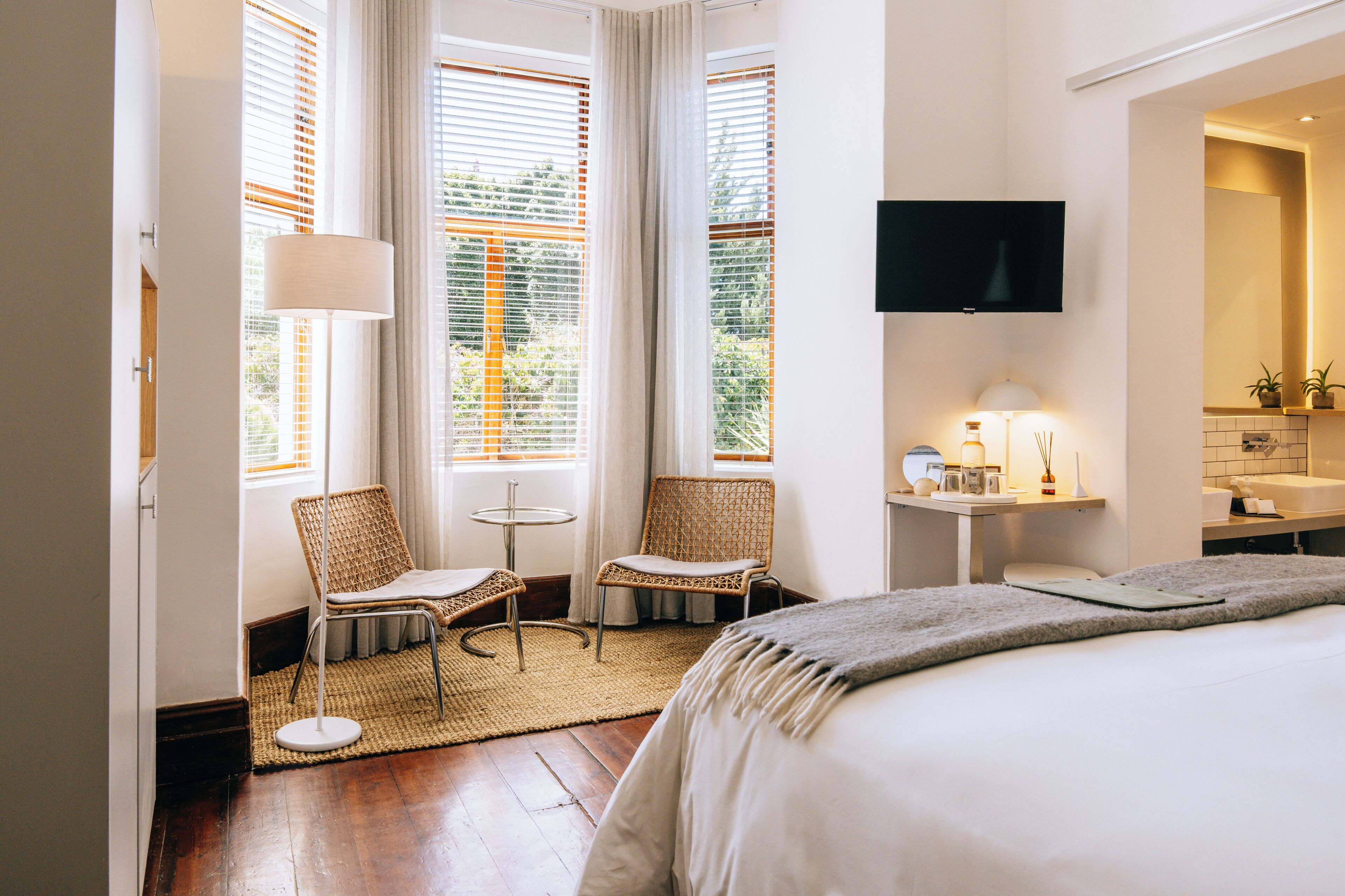 A hotel room with a bed, two wicker chairs, a small table, a TV, and a bathroom with a mirror and sink.