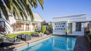 A modern pool area with a wooden deck, lounge chairs, and a white building with a glass door. - Cape Diem Lodge  (Cape Town)