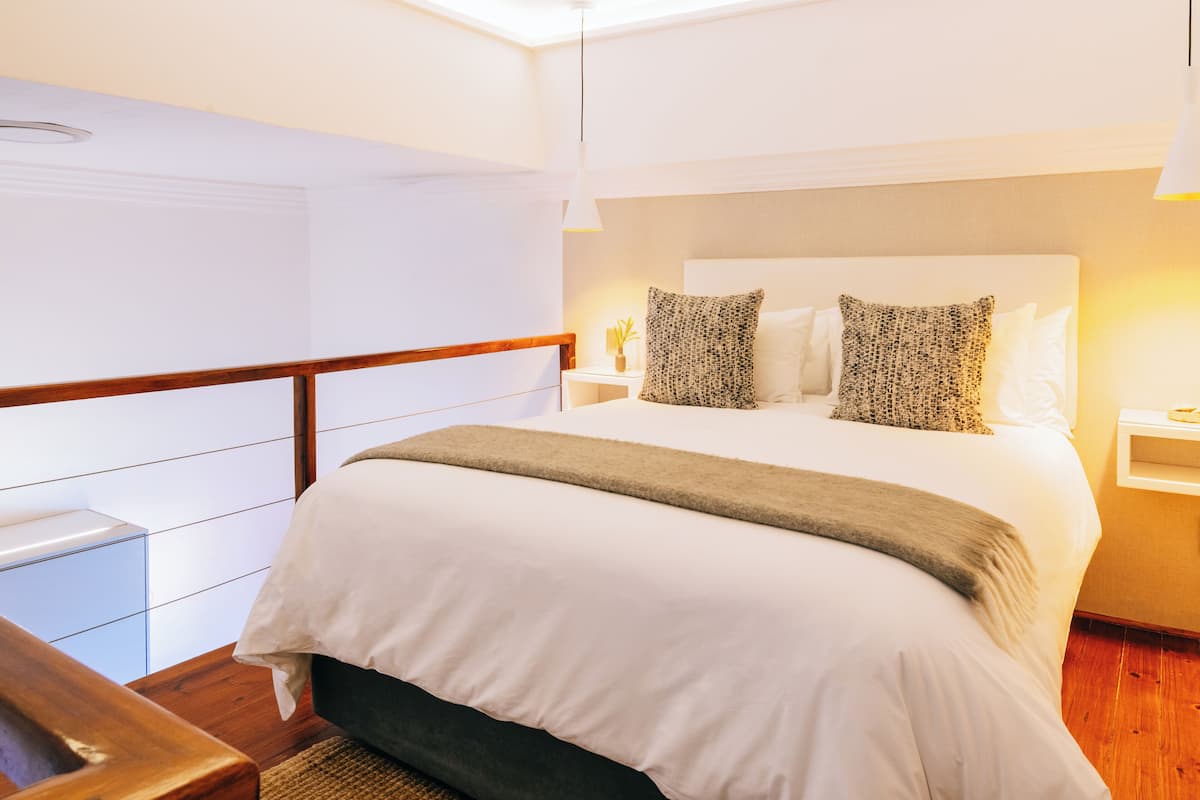 A neatly made bed with a white comforter and two patterned pillows, a bedside table with a lamp, and a wooden nightstand.