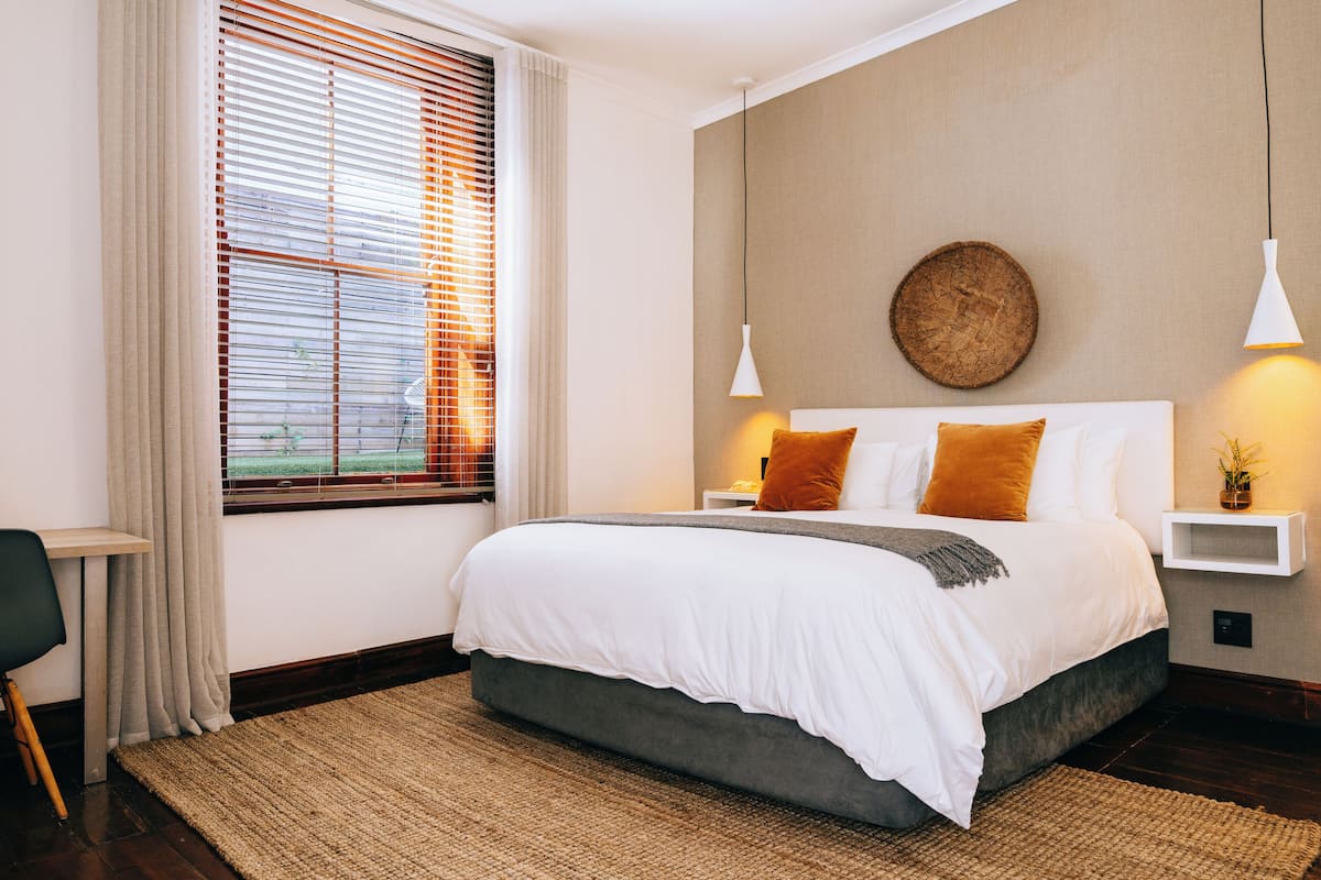 A modern bedroom with a large bed, a bedside table, a nightstand, a wall-mounted lamp, and a window with blinds.