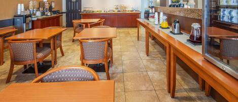 Zona de desayuno de hotel con mesas y sillas de madera, un mostrador de buffet y una estación de café.