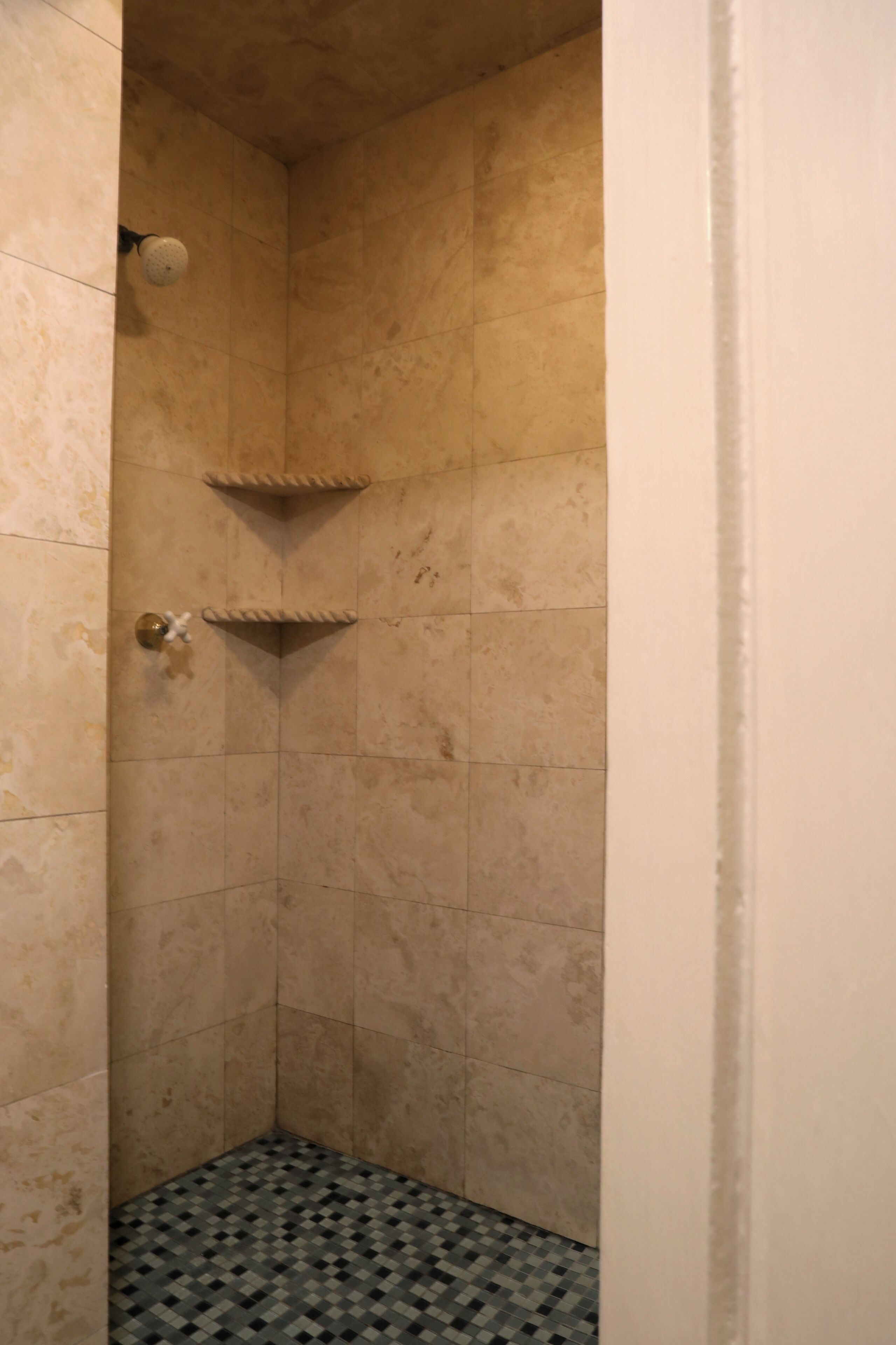 A corner shower with beige tiles, a small shelf, and a patterned floor.