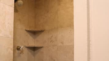 A corner shower with beige tiles, a small shelf, and a patterned floor.