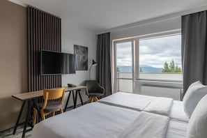 A hotel room with a bed, a desk, a chair, a TV, and a window with a view of the mountains.