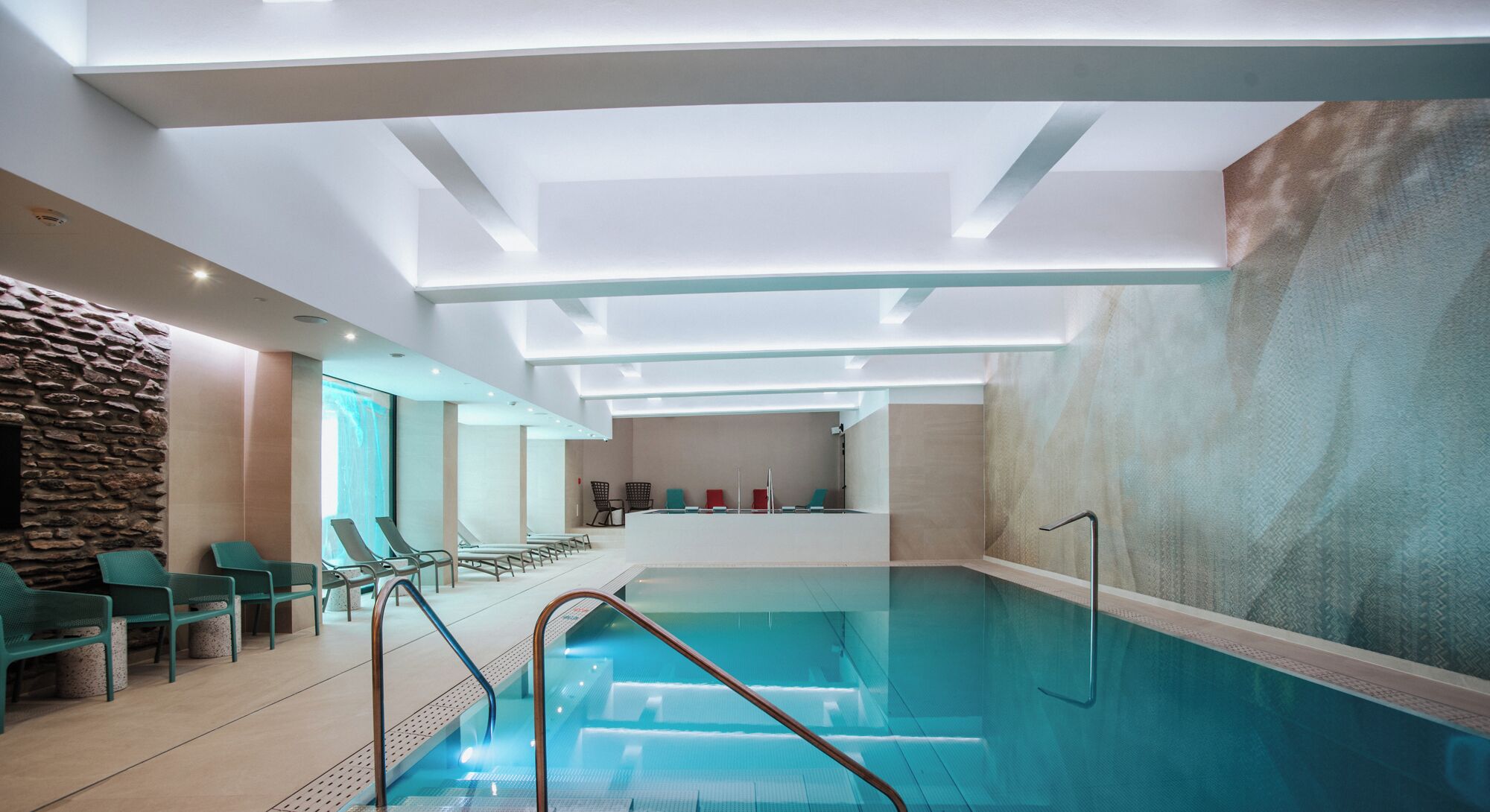 Une piscine intérieure moderne, avec de l’eau cristalline, un mur en pierre sur un côté, et un espace détente avec des chaises et un comptoir en arrière-plan.