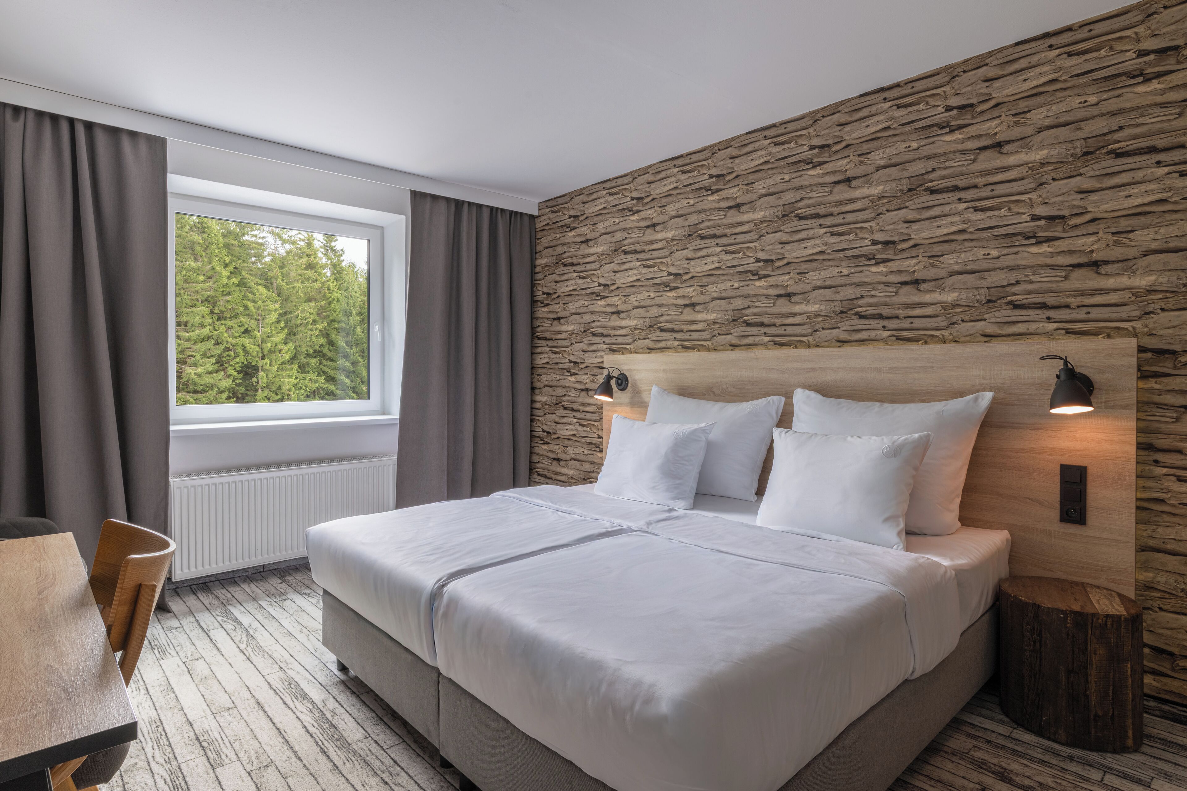 Une chambre d’hôtel moderne avec un grand lit, une tête de lit en bois, un mur décoratif en pierre, une fenêtre avec des rideaux et une petite table.
