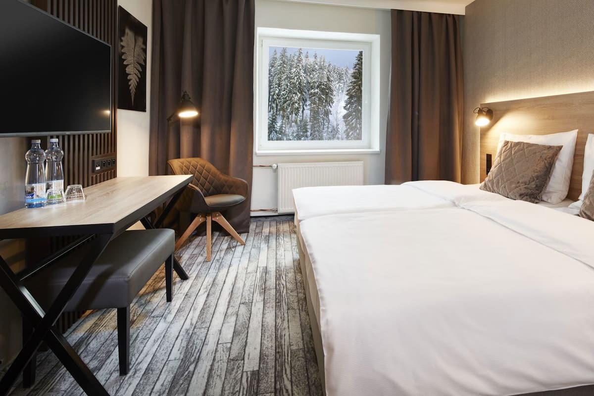 Habitación de hotel con cama, escritorio, silla, televisor y ventana con vista a árboles.