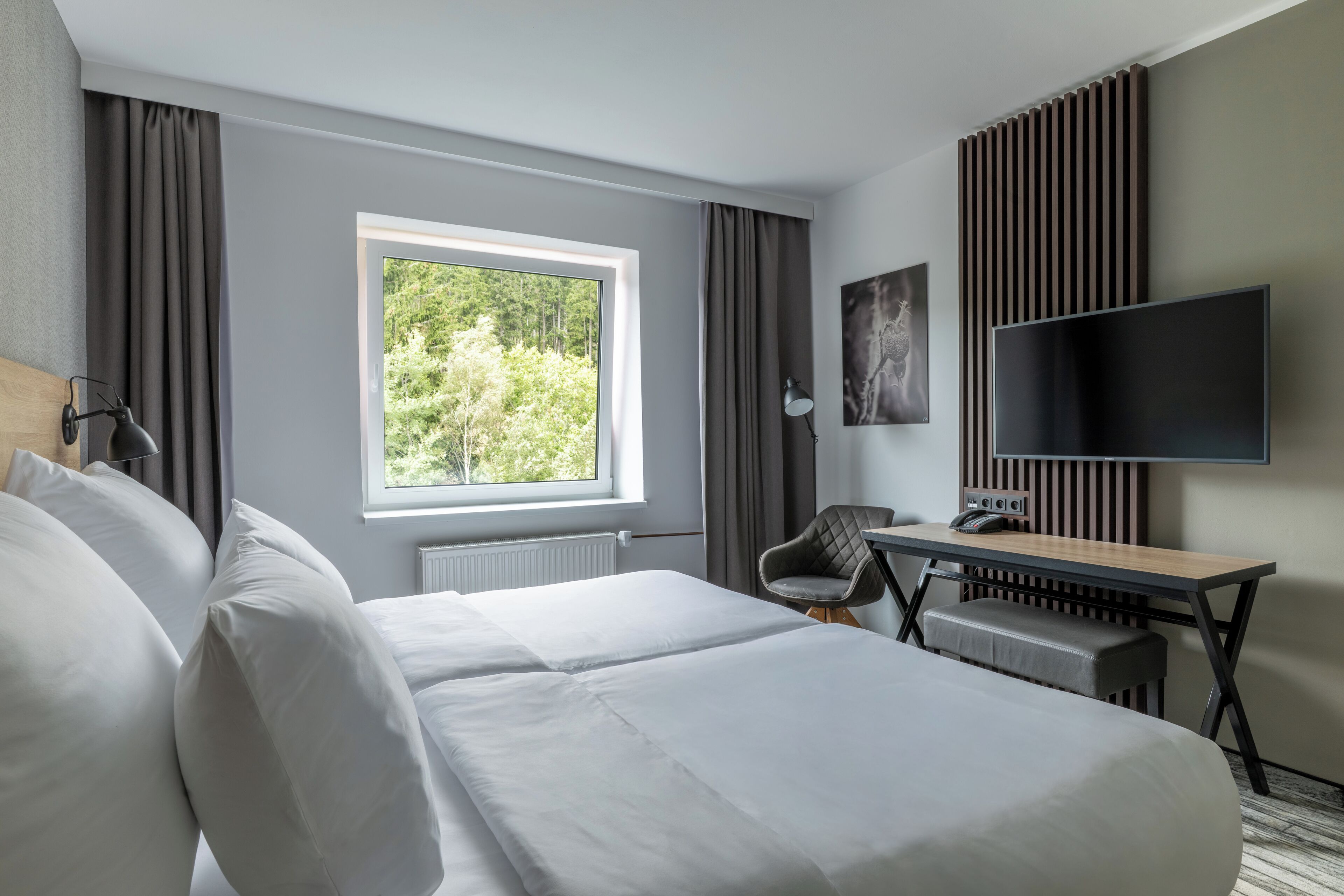 A modern hotel room with a large bed, a desk, a chair, a flat-screen TV, and a window with a view of greenery.