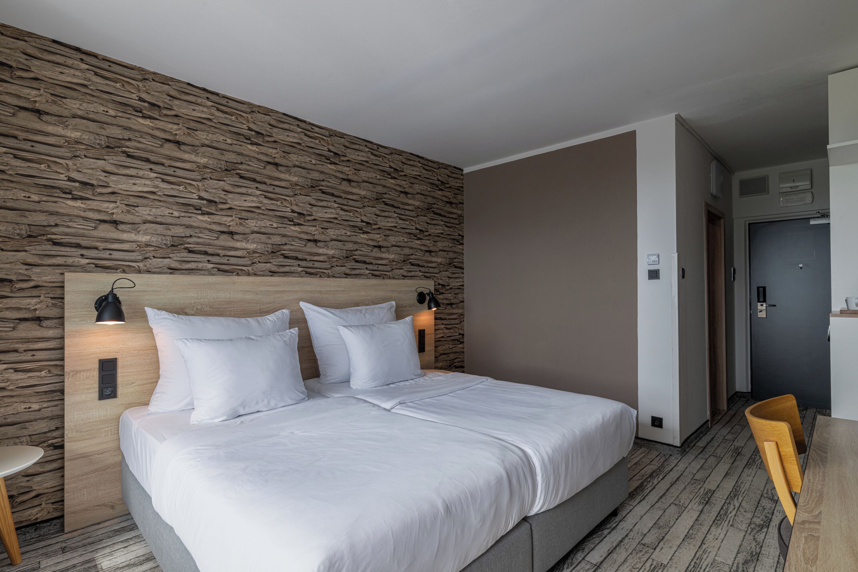 A modern hotel room with a stone-textured wall, a large bed with white bedding, two bedside lamps, and a wooden desk with a chair.