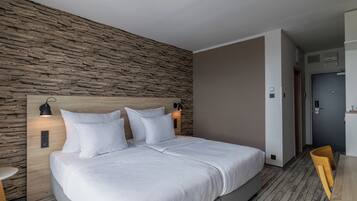 A modern hotel room with a stone-textured wall, a large bed with white bedding, two bedside lamps, and a wooden desk with a chair.
