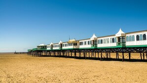 Una hilera de edificios blancos con techos verdes y balcones, construida sobre un muelle de madera que se adentra en una playa arenosa.