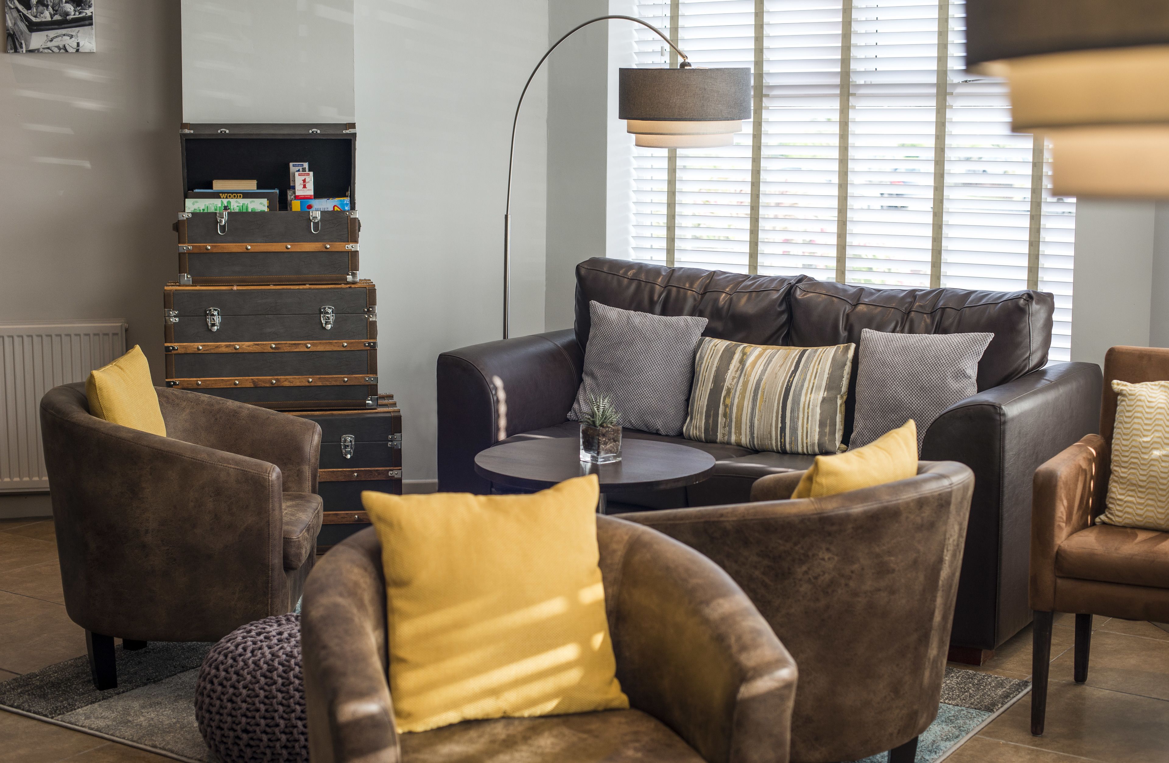 A living room with a leather sofa, armchairs, and a coffee table. There is a stack of suitcases against the wall and a floor lamp.