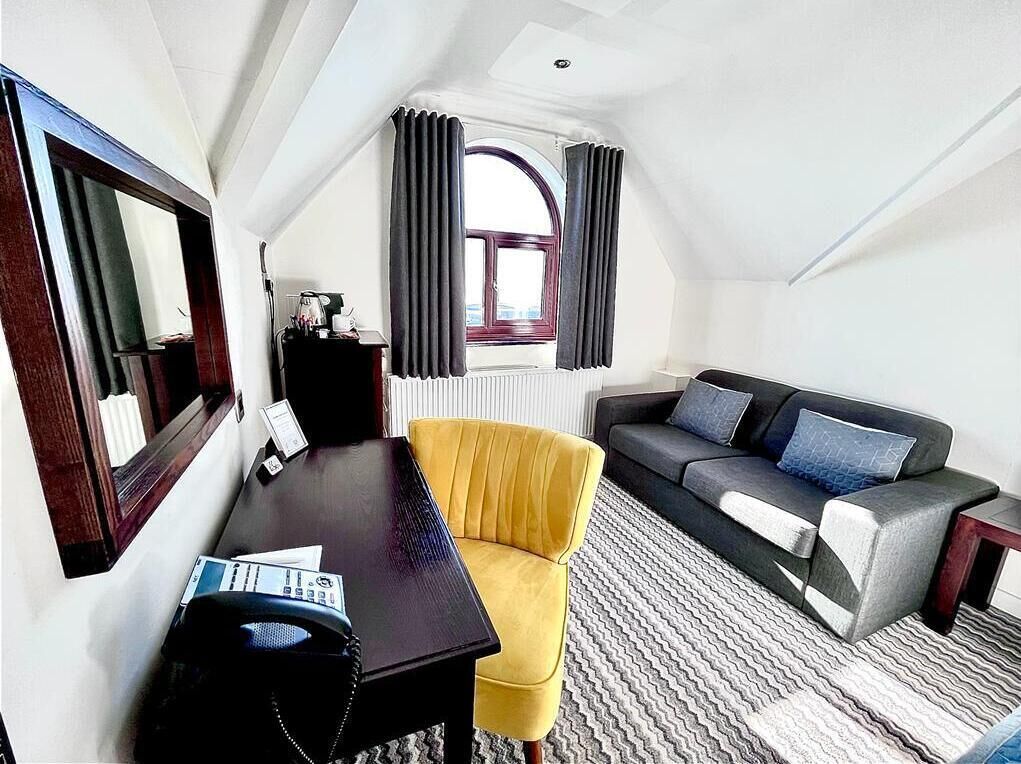 Quarto de hotel pequeno com uma escrivaninha de madeira escura, uma poltrona amarela, um sofá cinza com almofadas azuis e um espelho grande na parede esquerda.