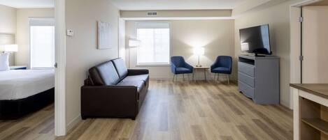 A modern hotel room with a bed, a brown leather sofa, two blue armchairs, a small table with a lamp, a TV on a cabinet, and a wooden floor.