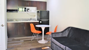 Cocina compacta con armarios de madera oscura, encimera blanca y refrigerador negro. Hay una mesa redonda blanca con dos sillas anaranjadas, y un sofá gris oscuro en la sala.