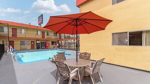 Un área de piscina con mesa y sillas, una sombrilla roja y un motel al fondo.