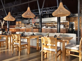 A restaurant interior with wooden tables and chairs, hanging wicker lamps, and a brick wall backdrop.