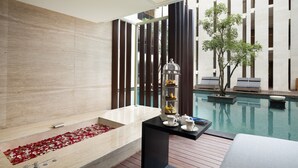 A modern bathroom with a large bathtub, a small table with cups and a decorative item, and a view of a pool with a wooden deck and a tree.