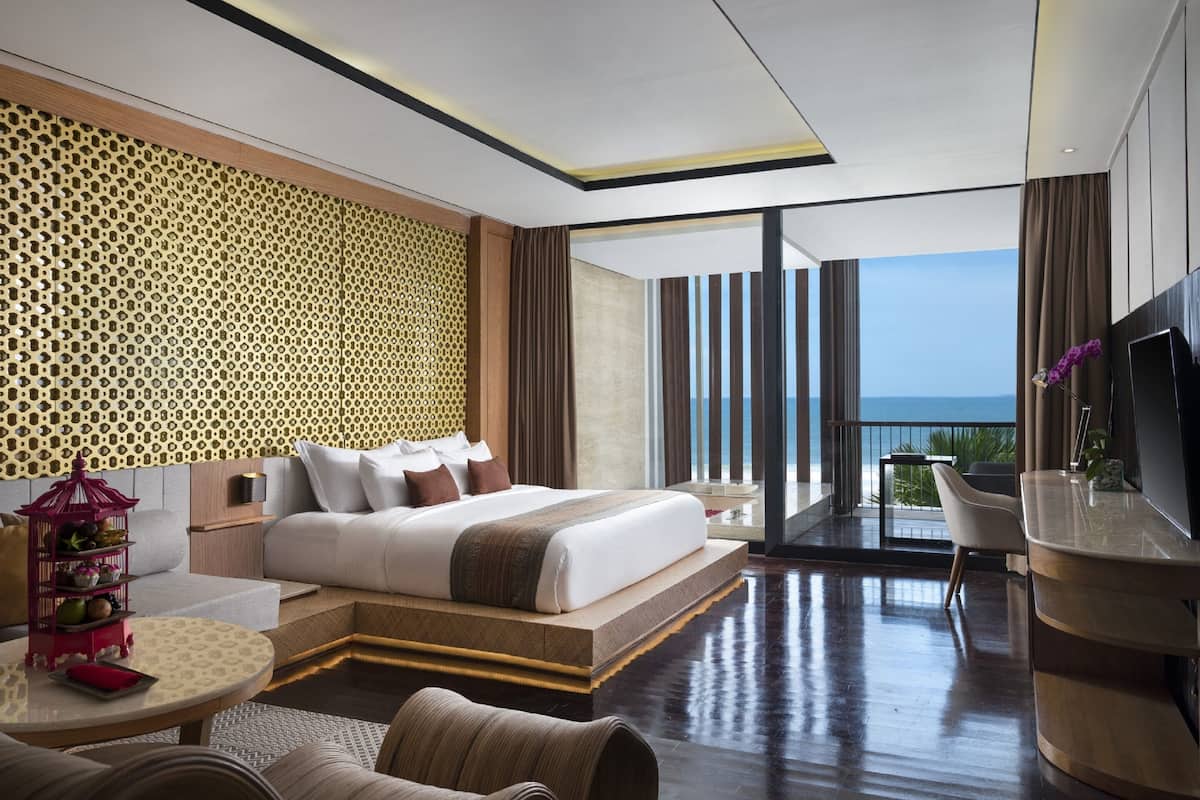 A modern hotel room with a large bed, a balcony with a view of the sea, and a dining area with a TV.