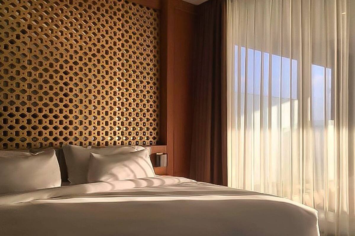 A hotel room with a large bed, a patterned headboard, a window with sheer curtains, and a view of the sky.