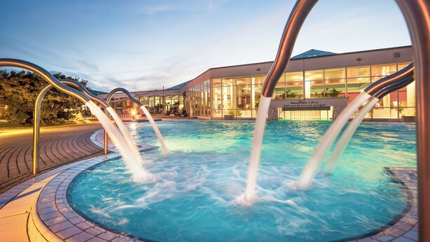 A swimming pool with multiple water jets, surrounded by a tiled deck and a modern building in the background. - Heide Spa Hotel & Resort (Bad Dueben)
