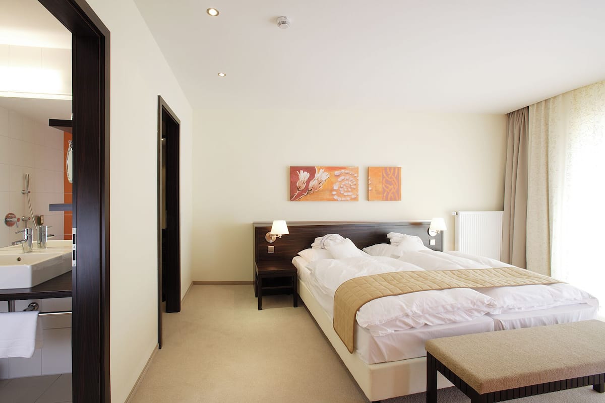 Habitación de hotel con cama, mesitas de noche, banco, espejo y baño con lavabo e inodoro.