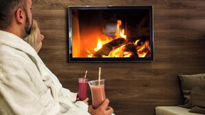 A person in a white bathrobe sitting on a couch, holding two drinks, with a fireplace and a TV mounted on a wooden wall. - Heide Spa Hotel & Resort (Bad Dueben)