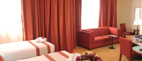 A hotel room with two beds, a red sofa, a dining table with a chair, and a fruit basket.