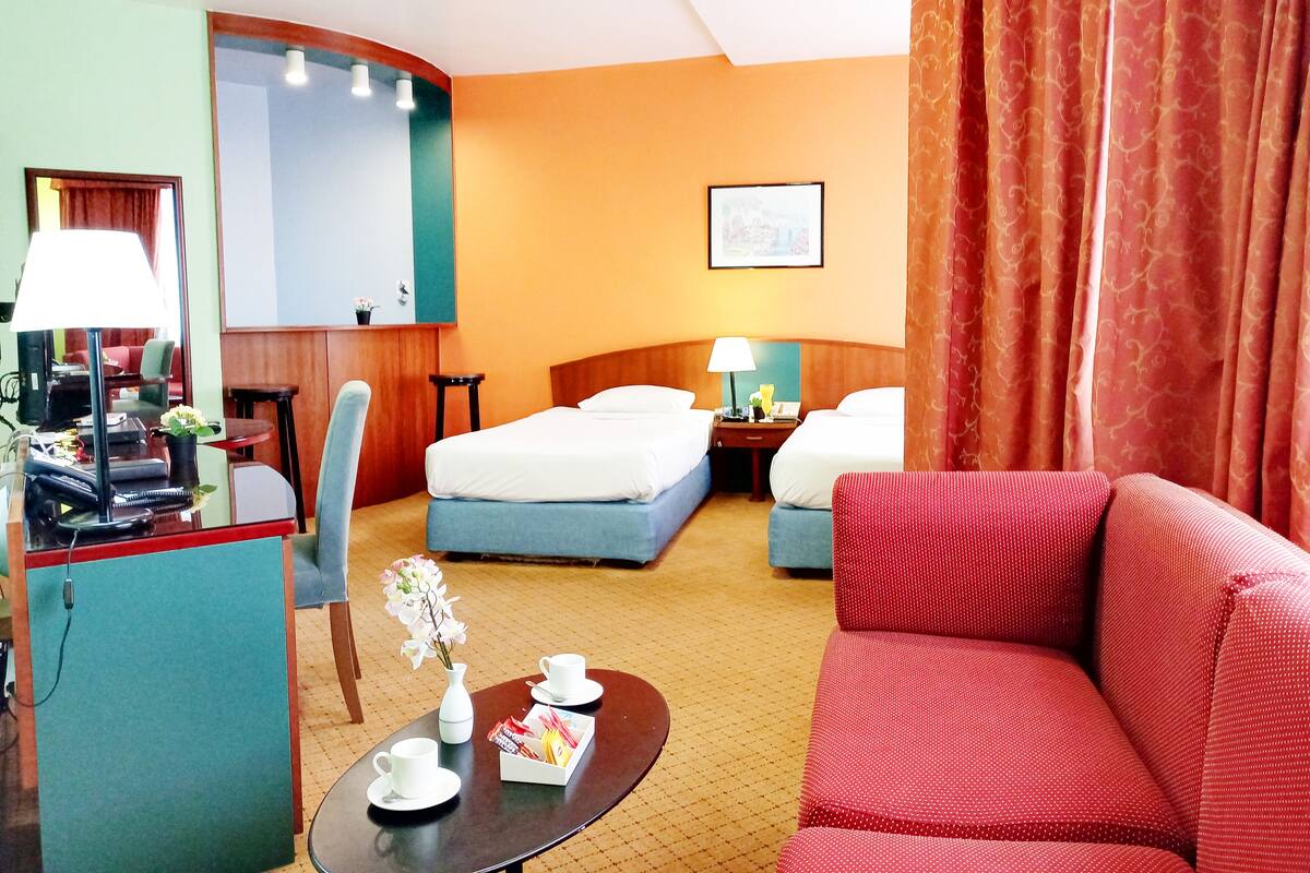 A hotel room with a bed, a desk with a lamp, a chair, a red sofa, and a small table with a vase and cups.