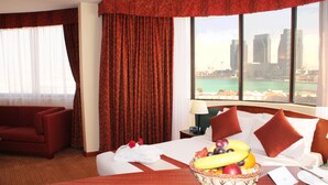 A hotel room with a bed, a red sofa, a fruit basket, and a city view through large windows.