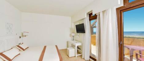 A hotel room with a bed, a desk with a TV, and a balcony with a view of the beach.