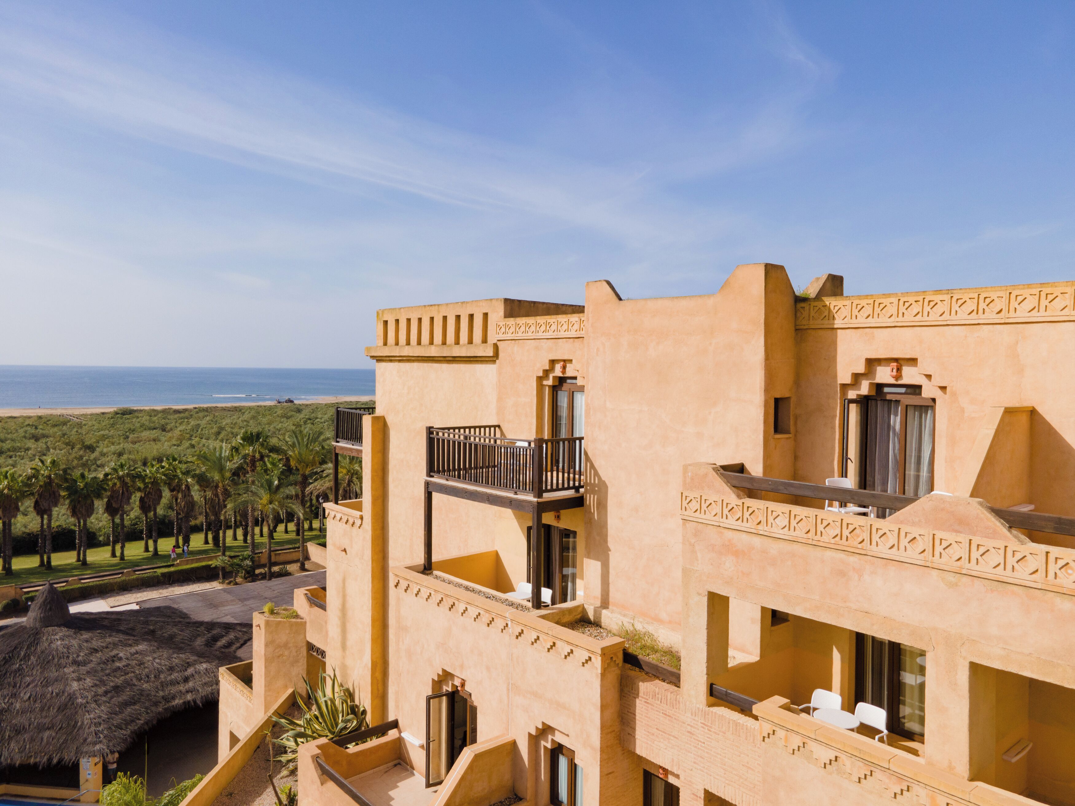 A multi-story building with balconies, a thatched-roof structure, and a view of the ocean.