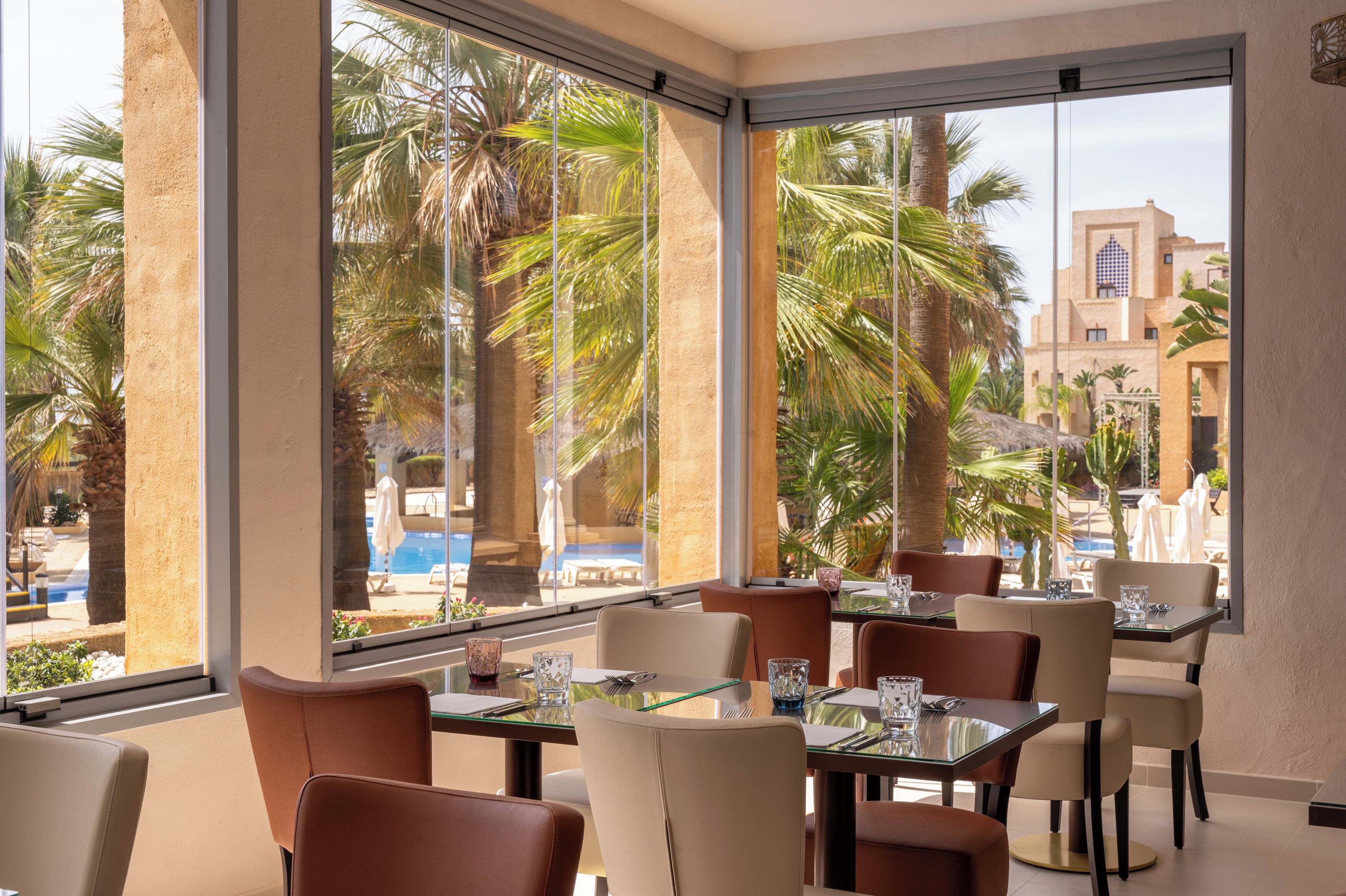 A hotel dining area with large windows, palm trees, and a view of a pool and building.