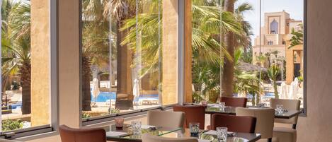 A hotel dining area with large windows, palm trees, and a view of a pool and building.