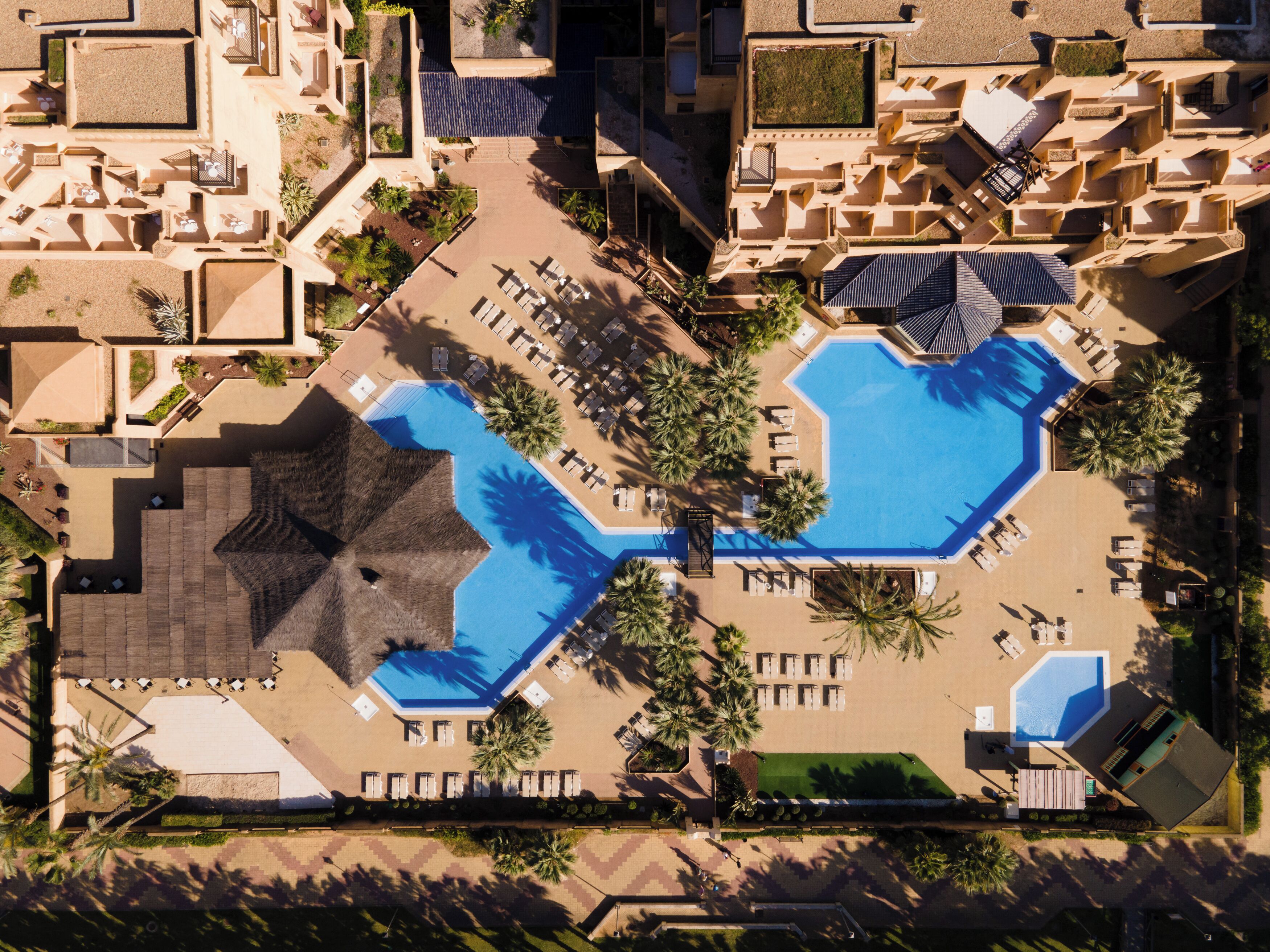 An aerial view of a resort with two swimming pools, lounge areas, and palm trees.