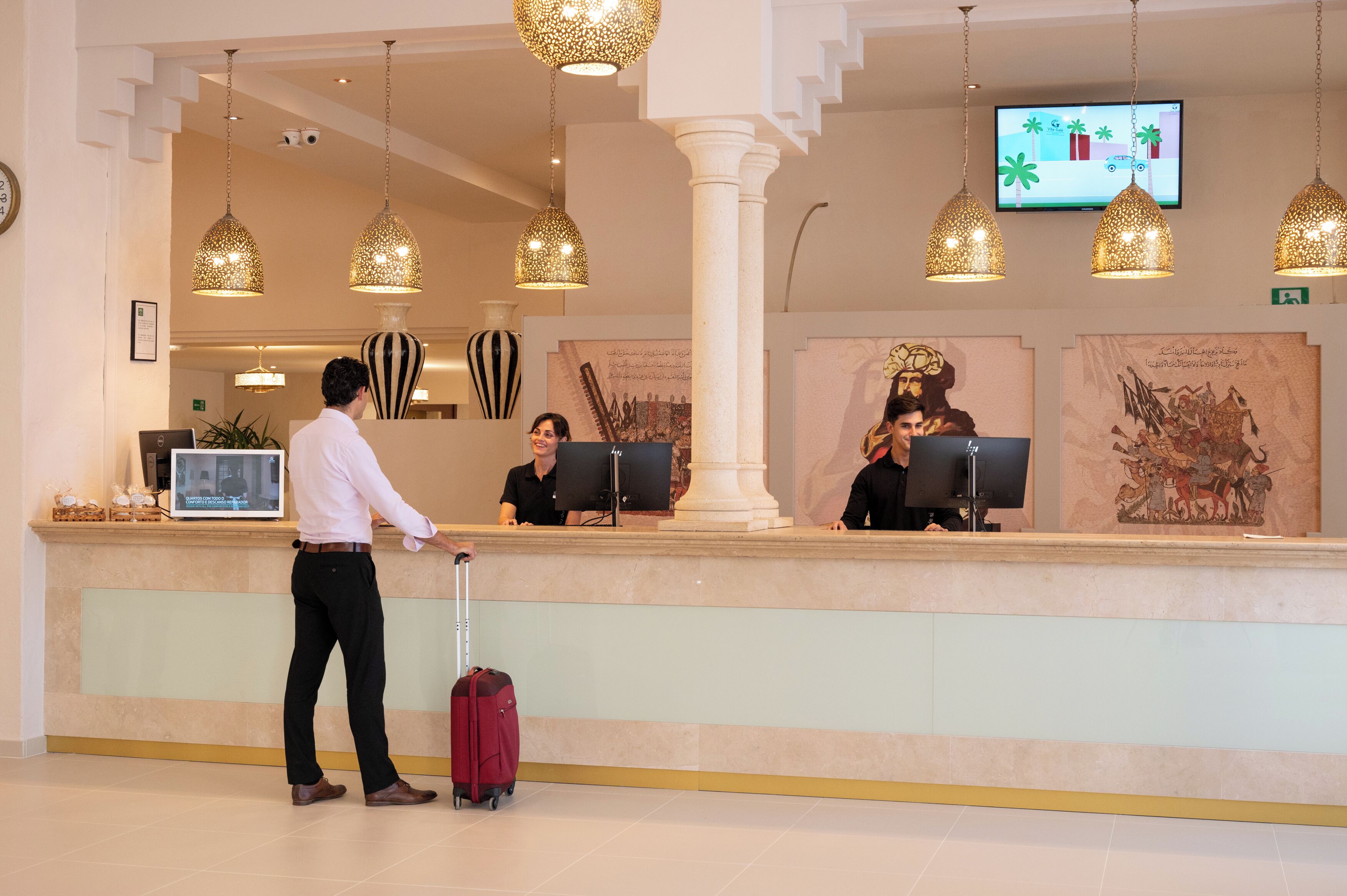 Un espace d’accueil d’hôtel comprenant un comptoir, des réceptionnistes, un téléviseur et un éclairage décoratif.