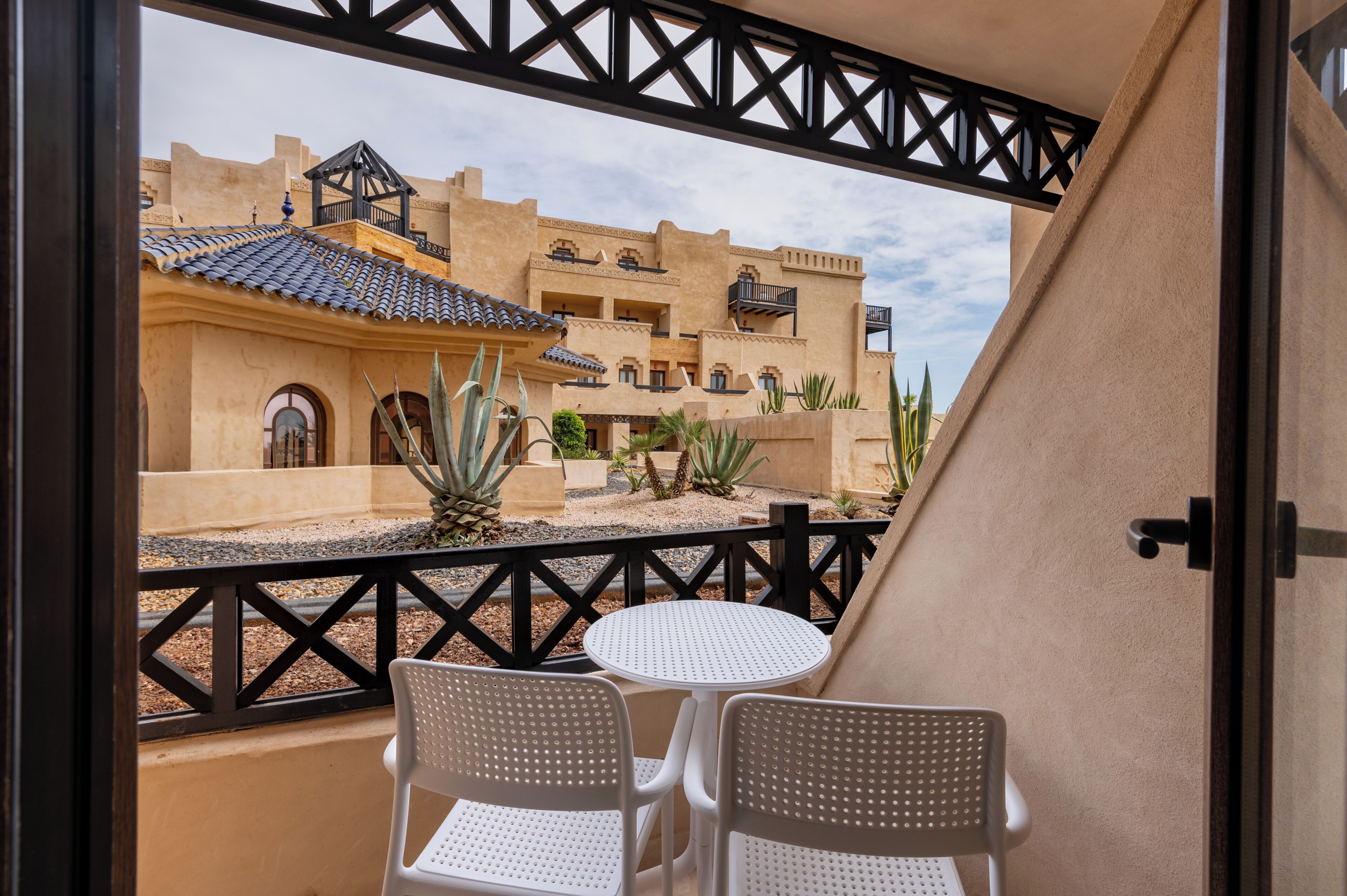 A balcony with two white chairs and a small round table, overlooking a building with a tiled roof and multiple balconies.