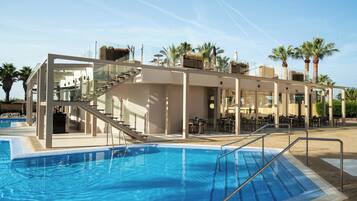 Una piscina con un deck con paredes de vidrio, mobiliario de exterior y palmeras.
