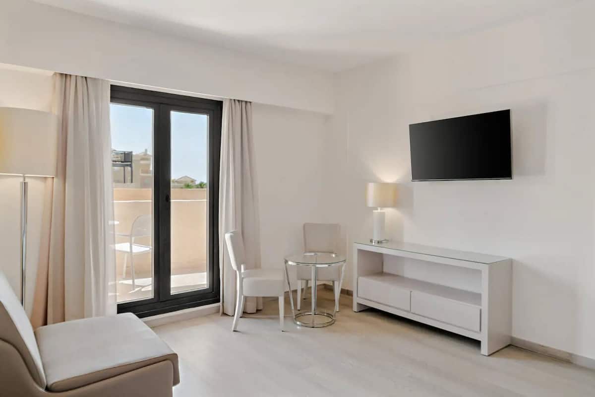 Ein modernes Hotelzimmer mit einer Couch, einem kleinen Tisch mit zwei Stühlen, einem an der Wand befestigten Fernseher und einer Schiebeglasdoor zu einem Außenbereich.