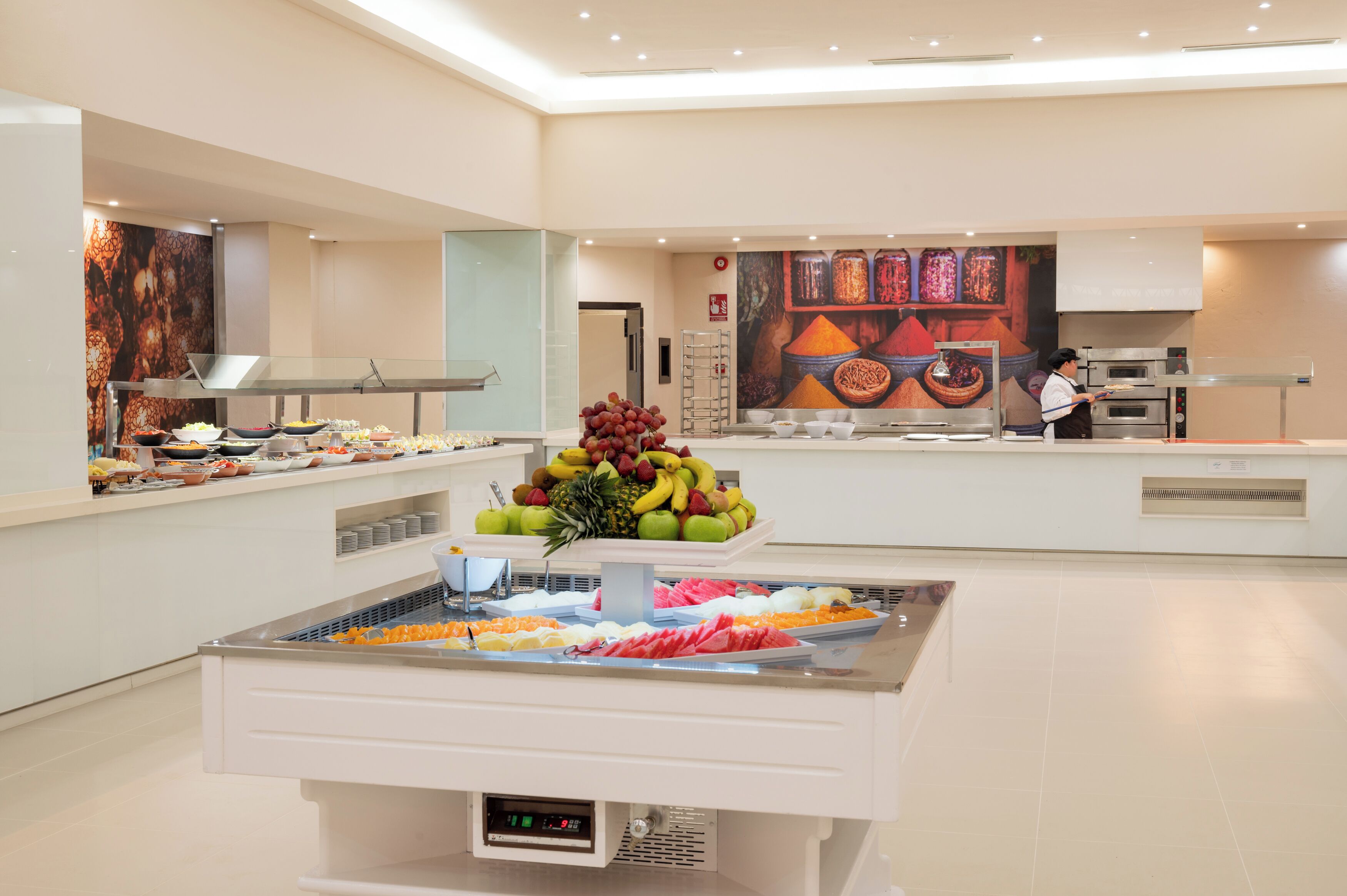 A buffet area with various food items, a fruit display, and a person in the background.