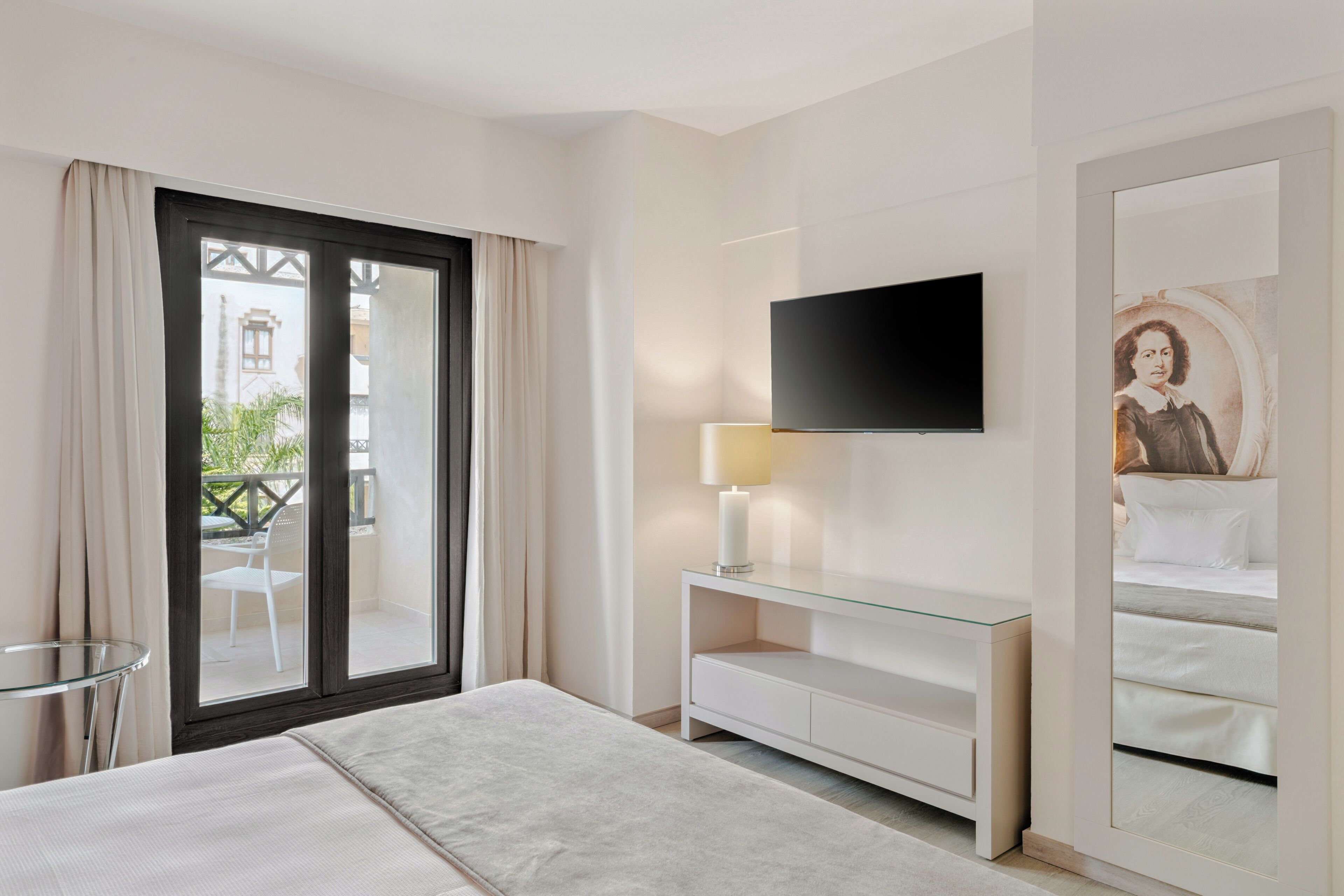 A modern hotel room with a bed, a TV mounted on the wall, a glass table, and a balcony with a view.