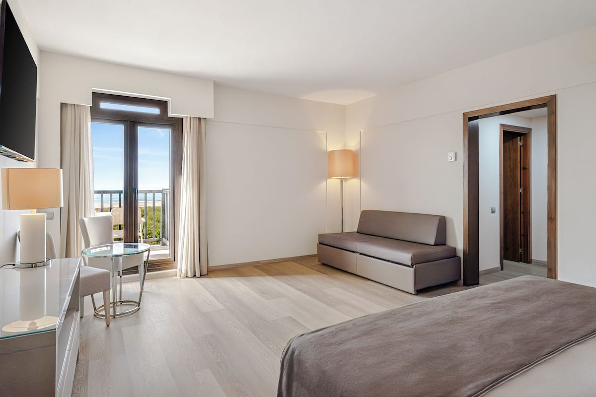 Ein modernes Hotelzimmer mit einem Bett, einer Couch, einem Glastisch mit Stühlen, einem Fernseher und einem Balkon mit Vorhängen.