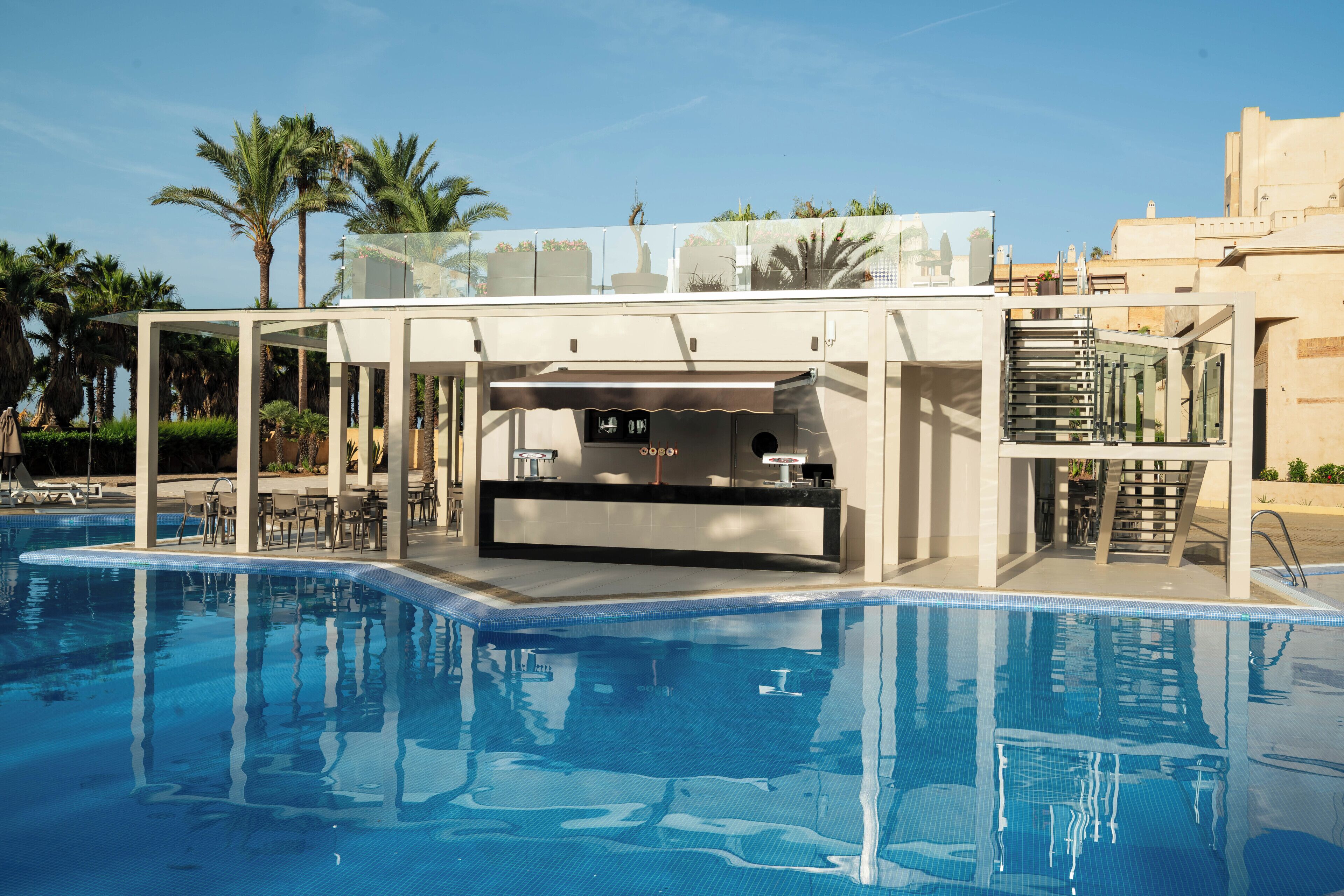 A pool area with a bar and seating, surrounded by palm trees and a clear sky.