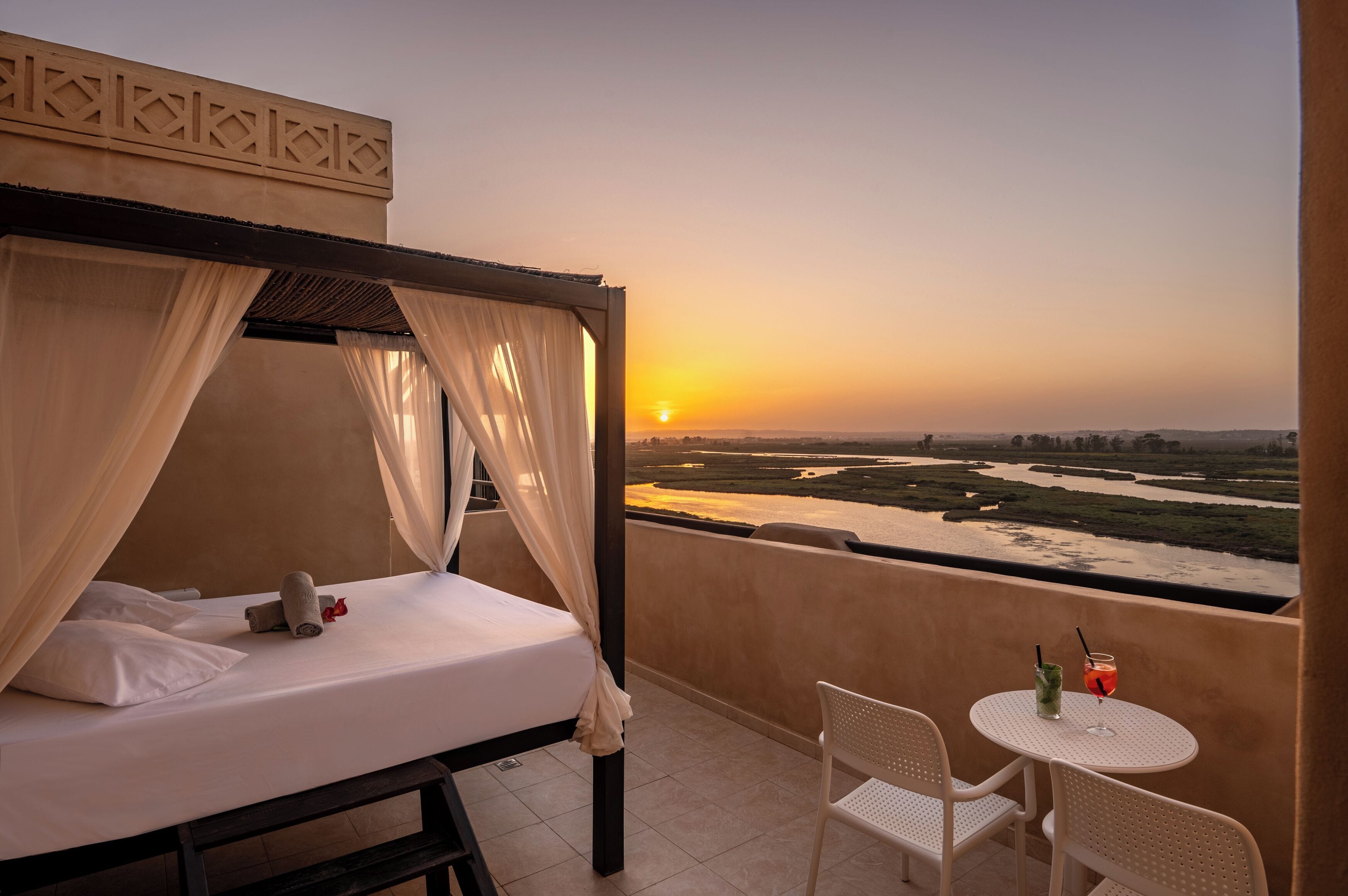 A balcony with a bed, a table with drinks, and a view of a river at sunset.