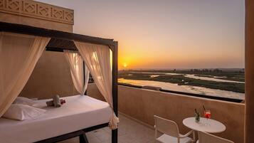 A balcony with a bed, a table with drinks, and a view of a river at sunset.