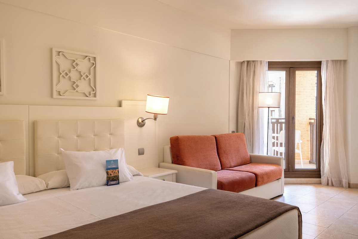 Ein Hotelzimmer mit Bett, Schlafsofa, Nachttisch, Wandlampe und Blick ins Freie durch eine Tür.